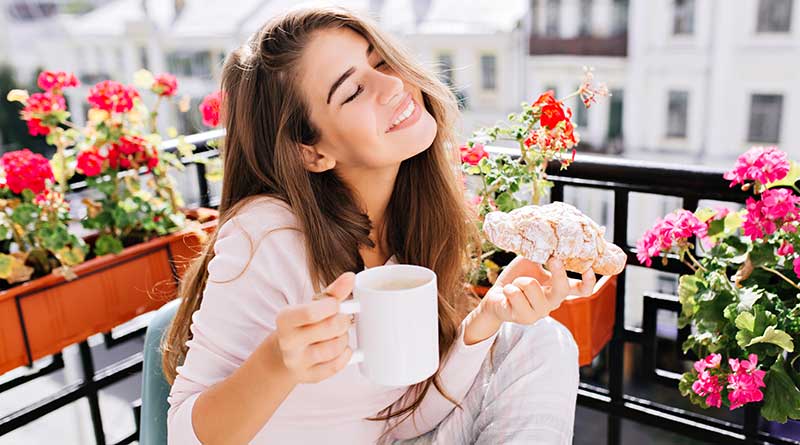 Happy young woman smiling and holding a mug of coffee and a croissant while sitting in her balcony